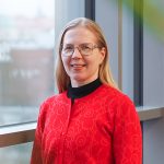 Kuvassa hymyilee Pirten työterveyshoitaja Sanna, jolla on pitkät vaaleat hiukset ja yllään punainen neuletakki.