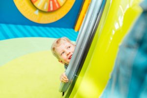 Kuvassa leikki-ikäinen vaaleatukkainen lapsi on piilosilla sairalaahuvipuiston värikkäässä ja leikintäyteisessä ympäristössä.