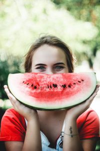 Kuvassa hymyilevä nainen, joka pitelee leveän hymyn muotoista vesimelonilohkoa hymynsä korkeudella.