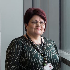 Kuvassa hymyilee Pirten työsuojelupäällikkö, hoitotyön päällikkö Hanna Peltoniemi. Hannalla on lyhyet punaiset hiukset ja silmälasit.