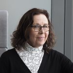 Kuvassa hymyilee Pirten työterveyshoitaja Outi, jolla on tummat olkapäille ulottuvat hiukset ja silmälasit. Outi ottaa vastaan työterveysasiakkaita Pirte Ratinassa.