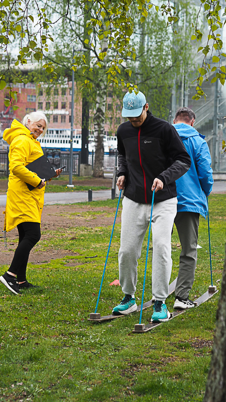 Kuvassa kaksi Korja-Kumin työntekijää kokeilevat tandemhiihtoa. Taustalla hymyilee Pirten työfysioterapeutti Kirsi keltaisessa takissa.