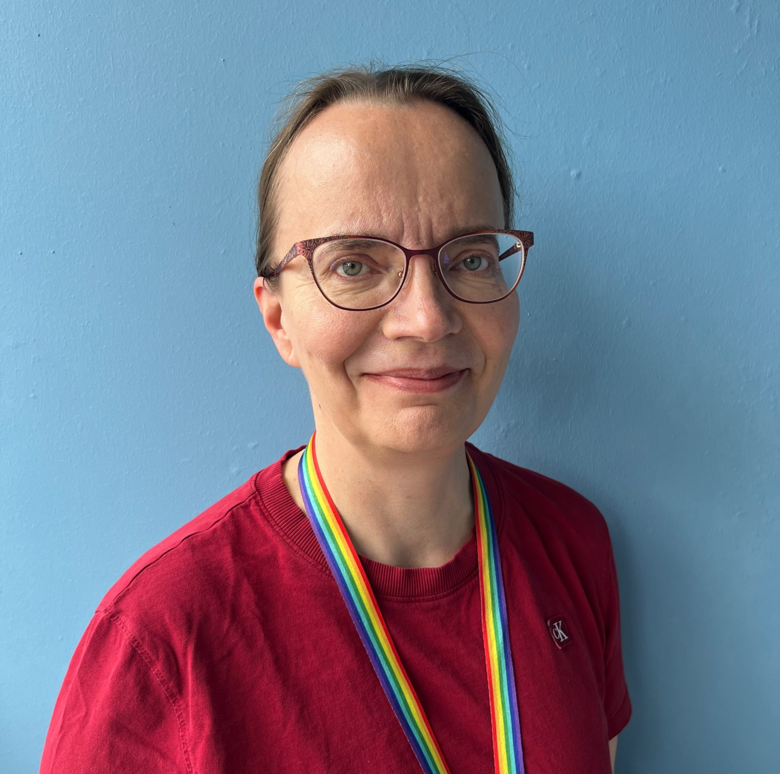 Kuvassa hymyilee Pirten lääkäri Marjut. Marjutilla on yllään silmälasit, punainen paita ja sateenkaariavainnauha. Taustalla näkyy vaaleansininen seinä.
